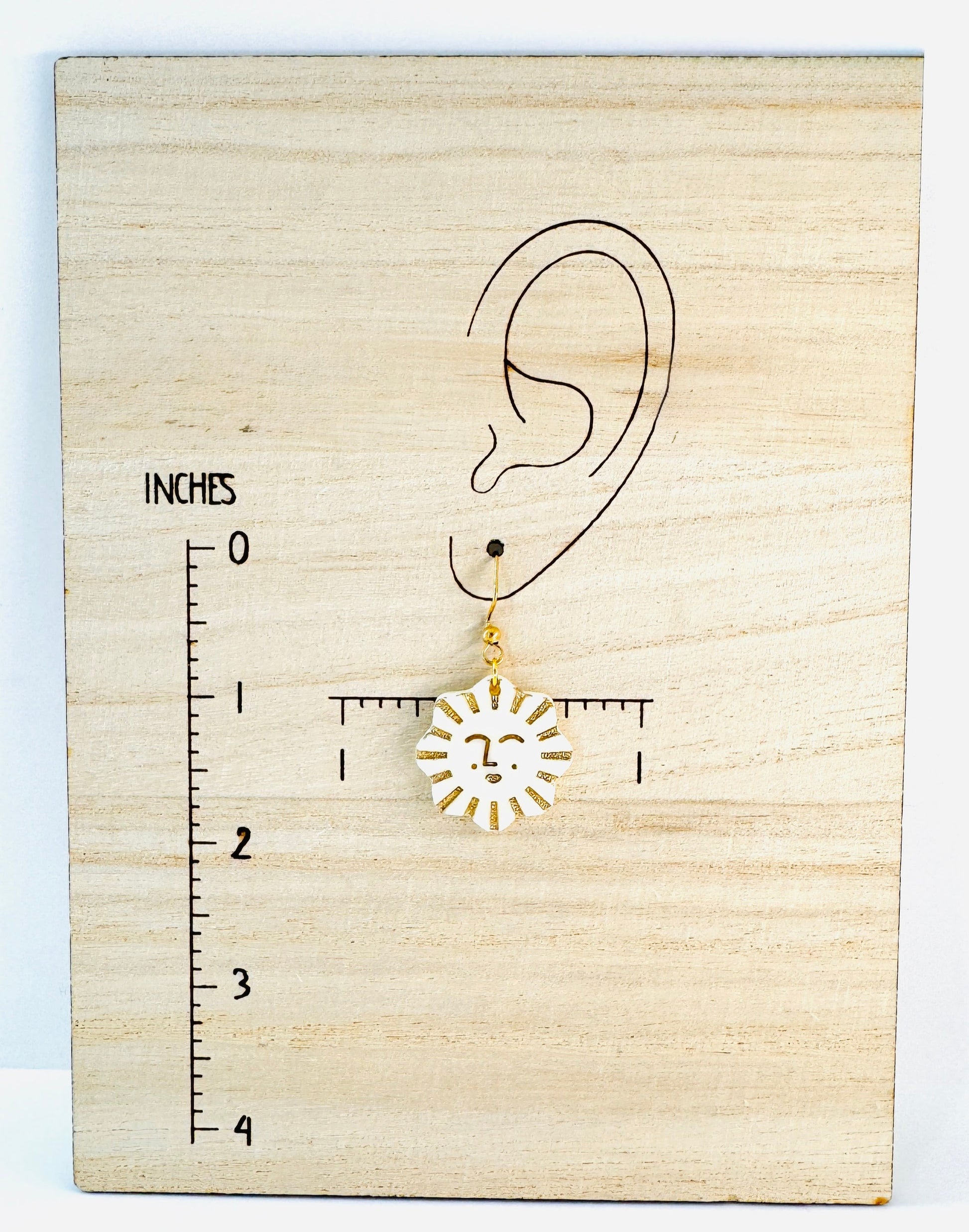 Gold sun-shaped earring on a wooden block with an ear illustration and ruler for scale.