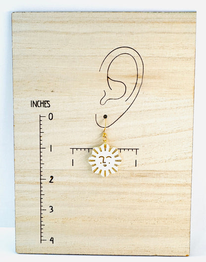 Gold sun-shaped earring on a wooden block with an ear illustration and ruler for scale.