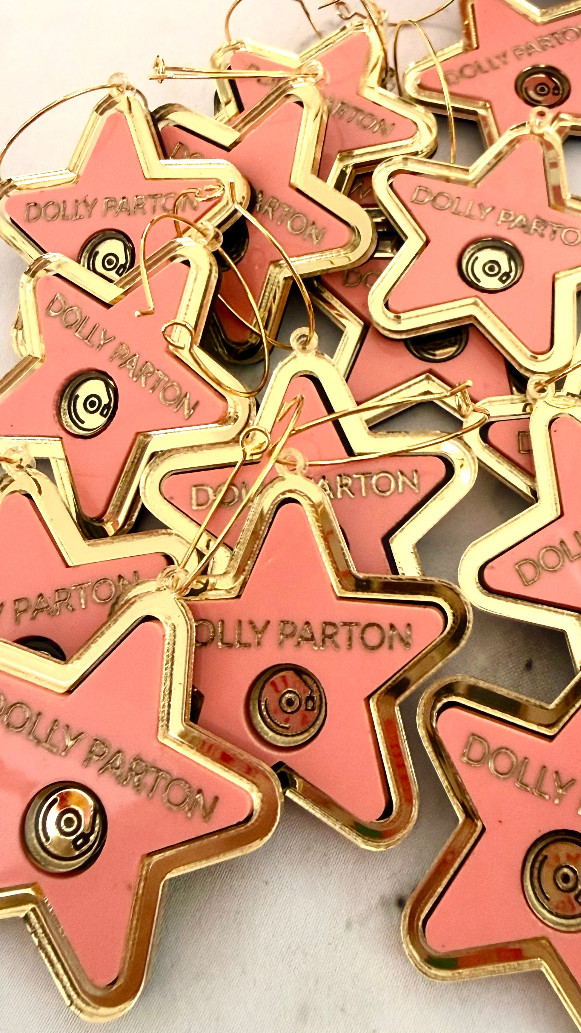 A pile of pink star-shaped earrings with gold metal borders and hangers, each engraved with the name 'DOLLY PARTON'.