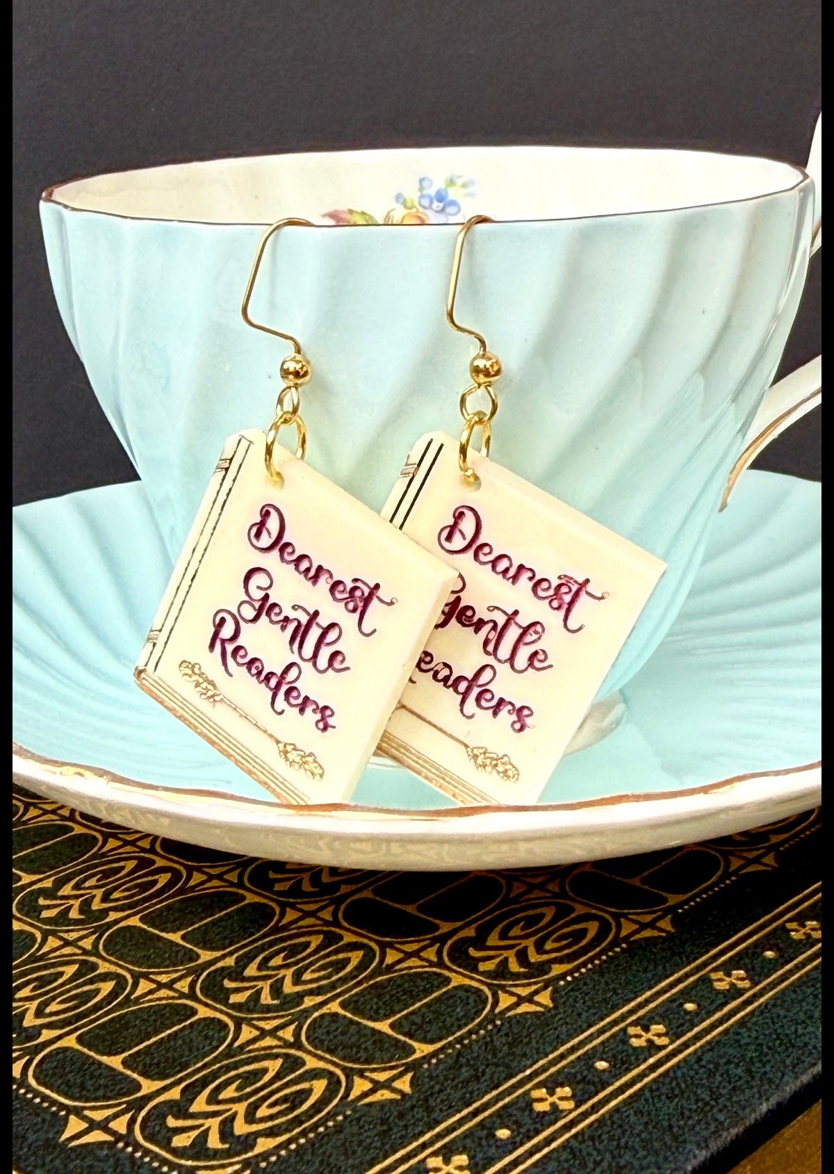 A pair of gold-tone dangle earrings with cream square charms printed with the phrase 'Dearest Gentle Readers' in maroon script, hanging from a light blue teacup on a matching saucer.