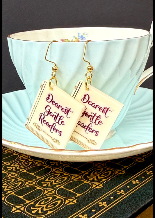 A pair of gold-tone dangle earrings with cream square charms printed with the phrase 'Dearest Gentle Readers' in maroon script, hanging from a light blue teacup on a matching saucer.