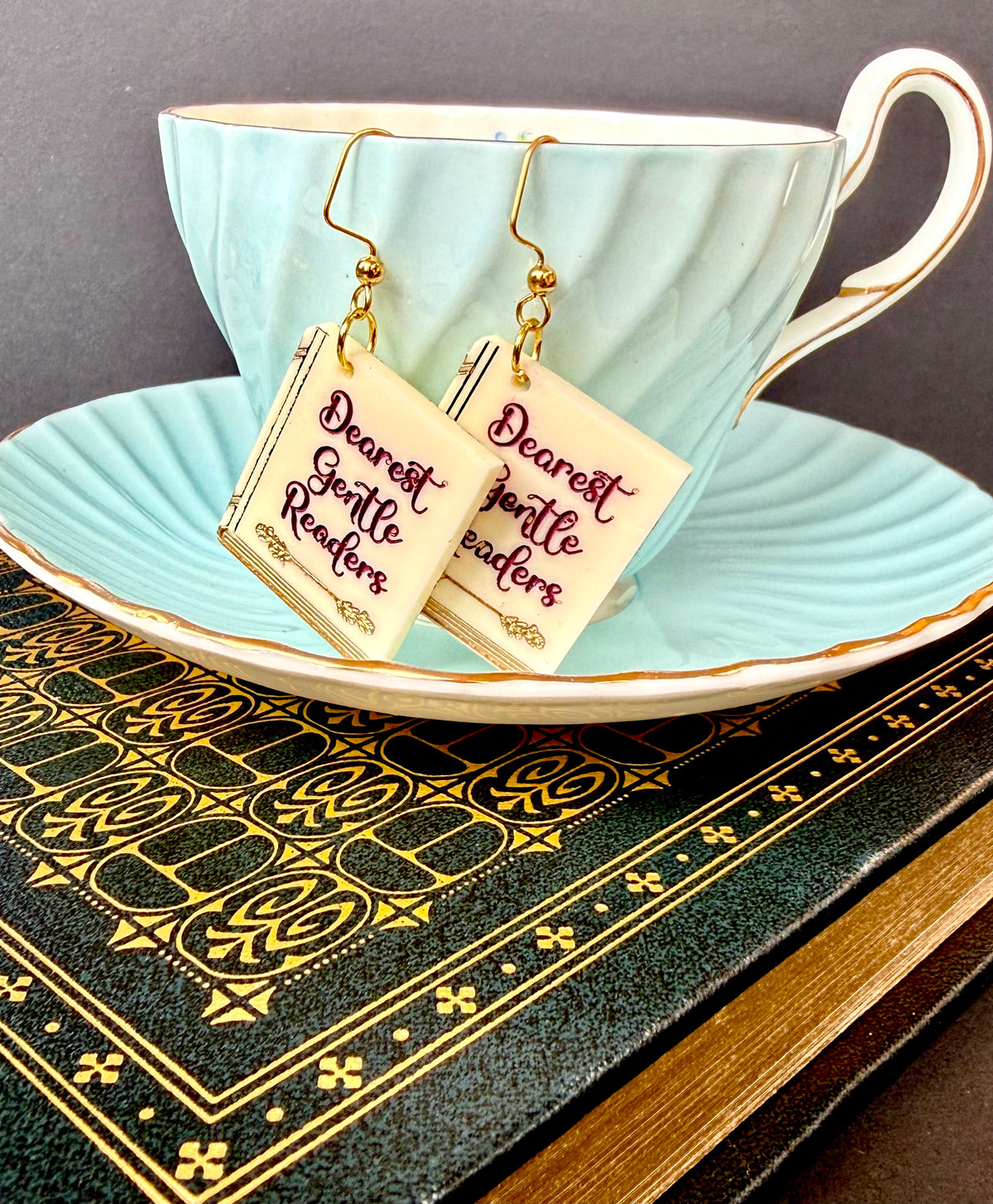 A pair of dangling earrings shaped like miniature books with gold hooks and cream-colored covers, printed with the phrase 'Dearest Gentle Readers' in dark red script. The earrings are displayed on a light blue teacup and saucer, resting on a dark green and gold patterned book against a dark gray background.