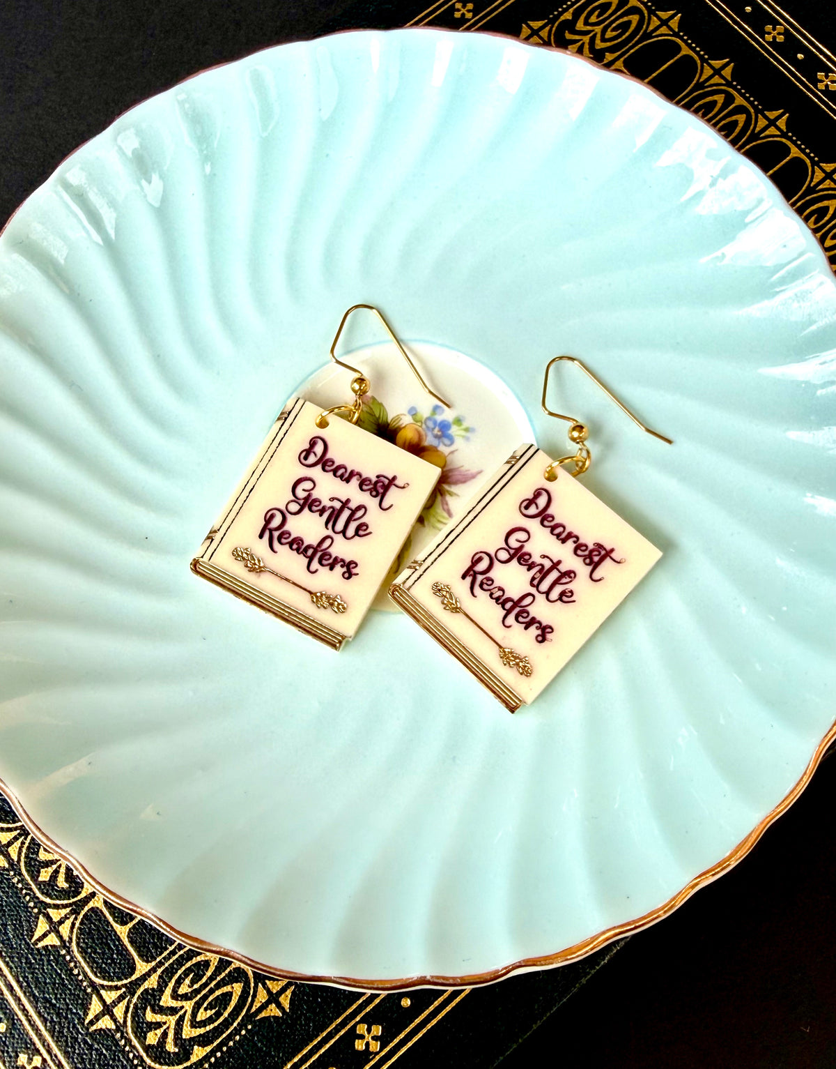 A pair of rectangular drop earrings shaped like miniature books with gold hooks, featuring the phrase 'Dearest Gentle Readers' in dark red script on a cream background, displayed on a light blue scalloped plate.