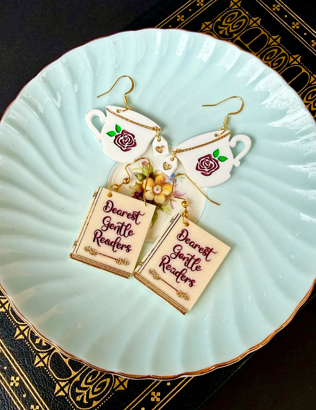 A pair of dangling earrings displayed on a pale blue plate. Each earring features a white teacup with a purple rose and gold-tone wire hooks, with a dangling cream-colored book charm that reads 'Dearest Gentle Readers' in purple script.
