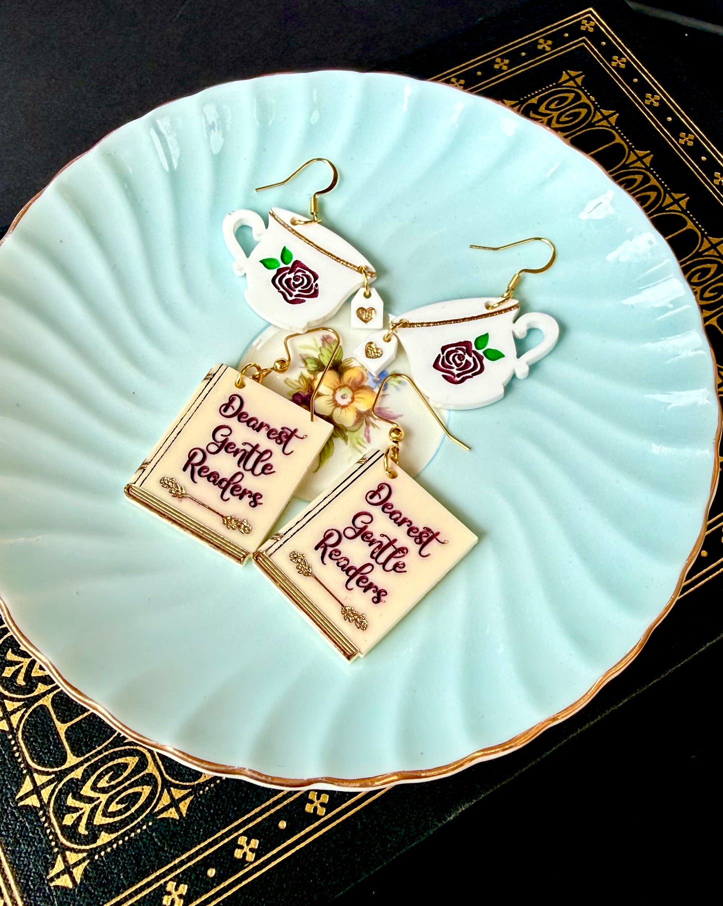 A pair of dangling earrings displayed on a light aqua ceramic plate. The earrings feature white teacups with pink rose details, gold hooks, and rectangular cream-colored book charms printed with the phrase 'Dearest Gentle Readers' in dark pink script, accented with small gold and pink floral elements.