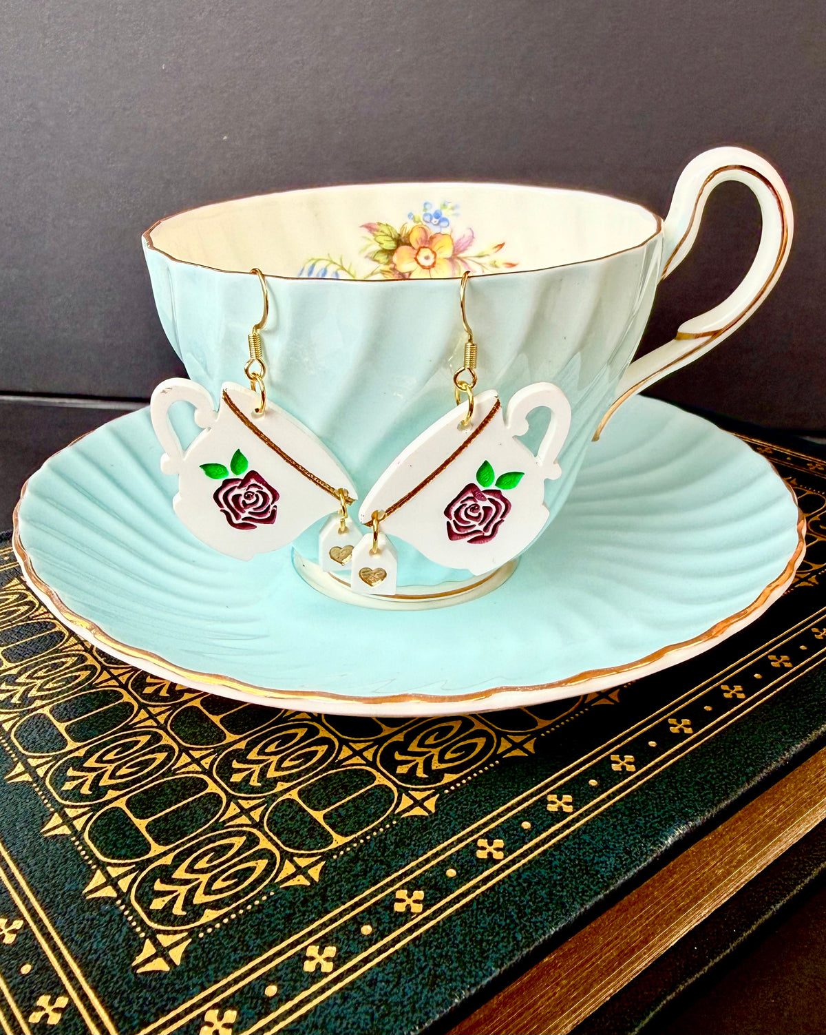 A pair of handmade earrings shaped like teacups with purple rose details, hanging from gold hooks and displayed on a light blue teacup and saucer set resting on a dark patterned book.