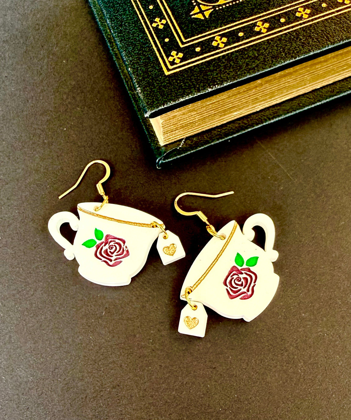 A pair of white teacup earrings with red rose and green leaf engravings, gold wire hooks, and small dangling gold tea bag charms, displayed on a dark brown surface next to an antique-style book.