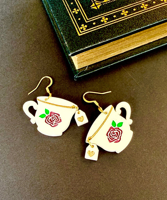 A pair of white teacup earrings with red rose and green leaf engravings, gold wire hooks, and small dangling gold tea bag charms, displayed on a dark brown surface next to an antique-style book.