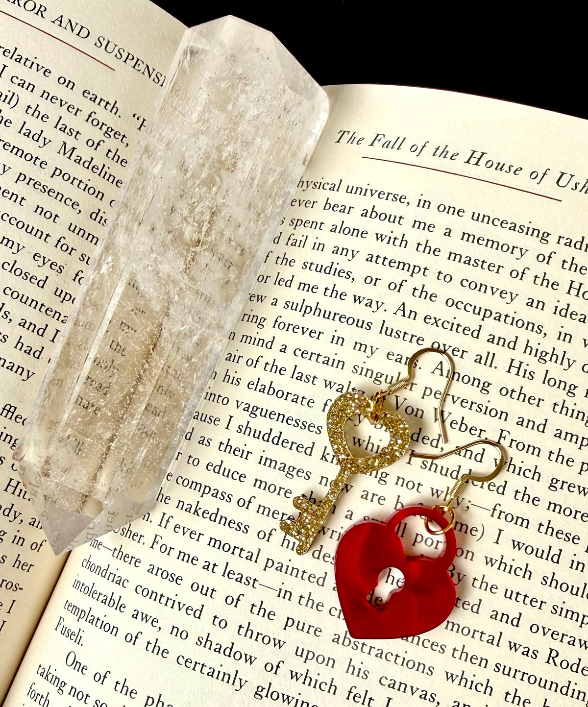A pair of mismatched earrings on a book page. One is a glittery golden key with a heart top, and the other is a heart shaped lock. The pair are finished with gold tone ear wires.