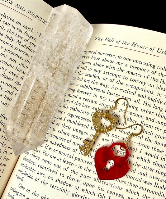 A pair of mismatched earrings on a book page. One is a glittery golden key with a heart top, and the other is a heart shaped lock. The pair are finished with gold tone ear wires.