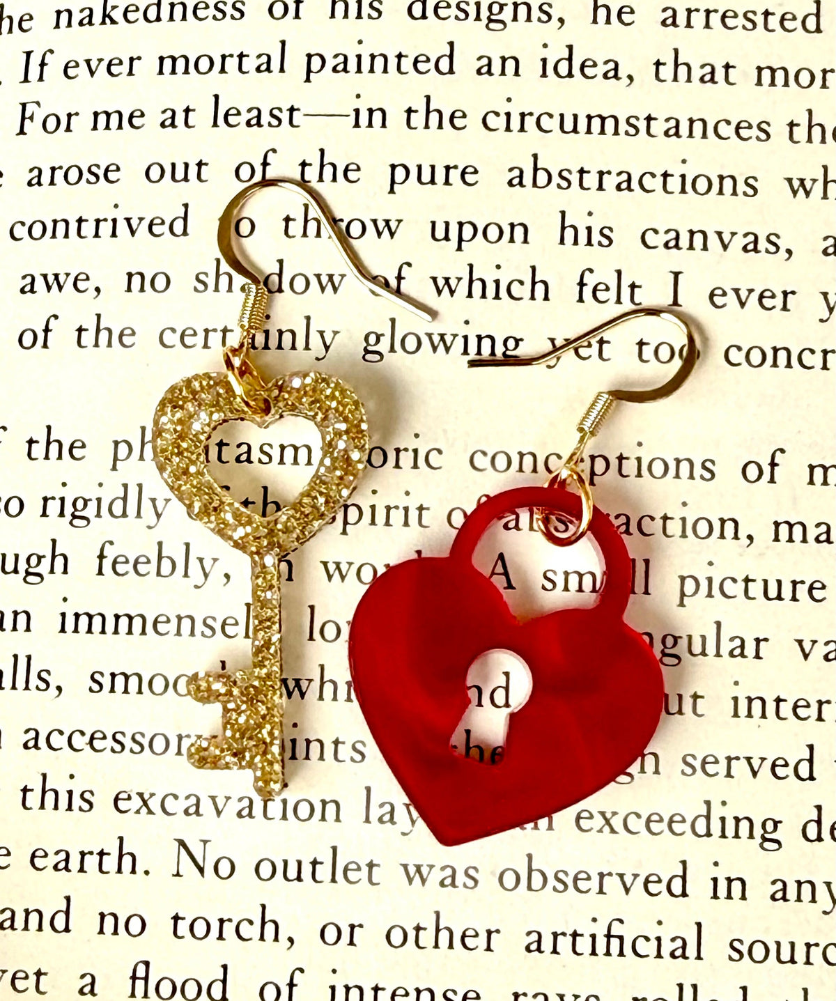 A pair of mismatched earrings on a book page. One is a glittery golden key with a heart top, and the other is a heart shaped lock. The pair are finished with gold tone ear wires.