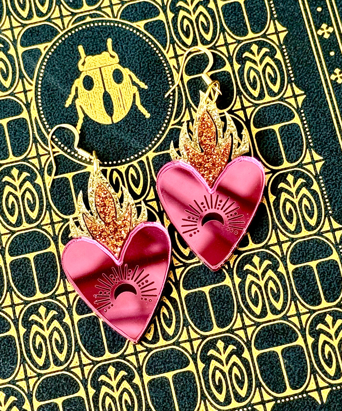 Pink celestial heart statement earrings with glitter flames displayed on a black and gold beetle patterned background