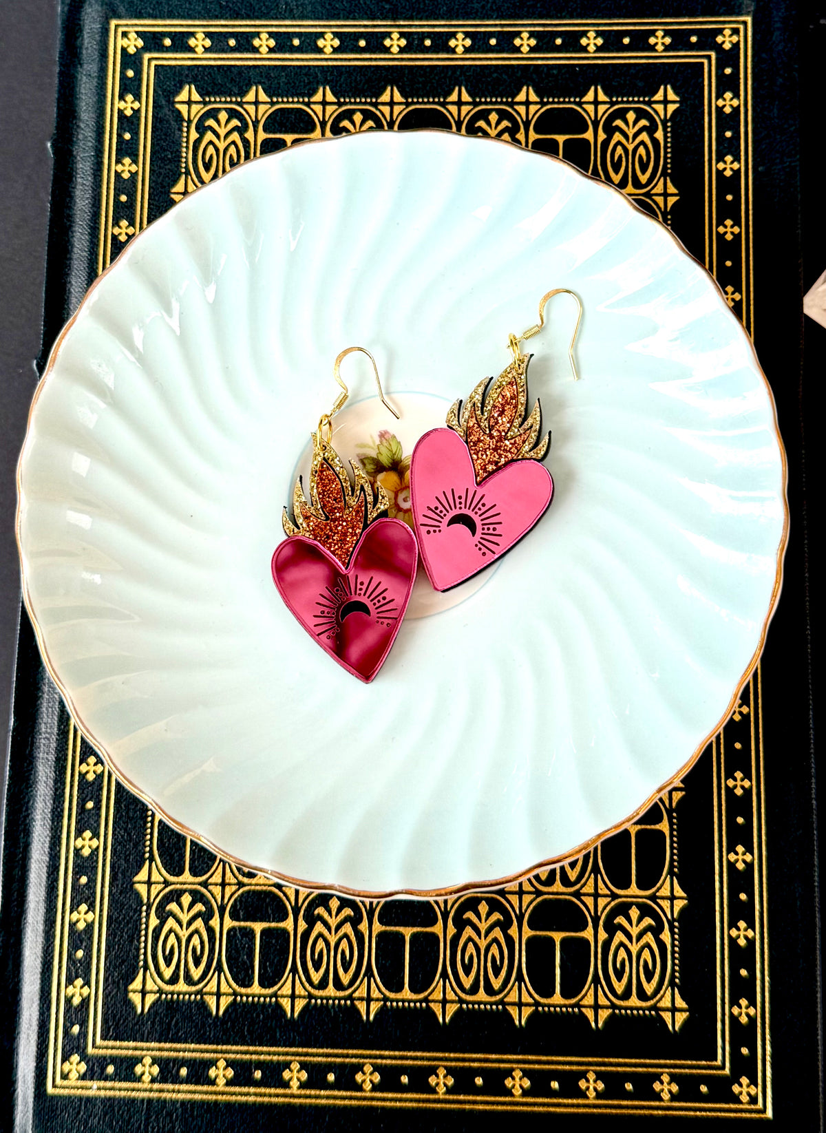 A pair of dangling heart earrings displayed on a white scalloped plate, set against a black book with ornate gold borders.
