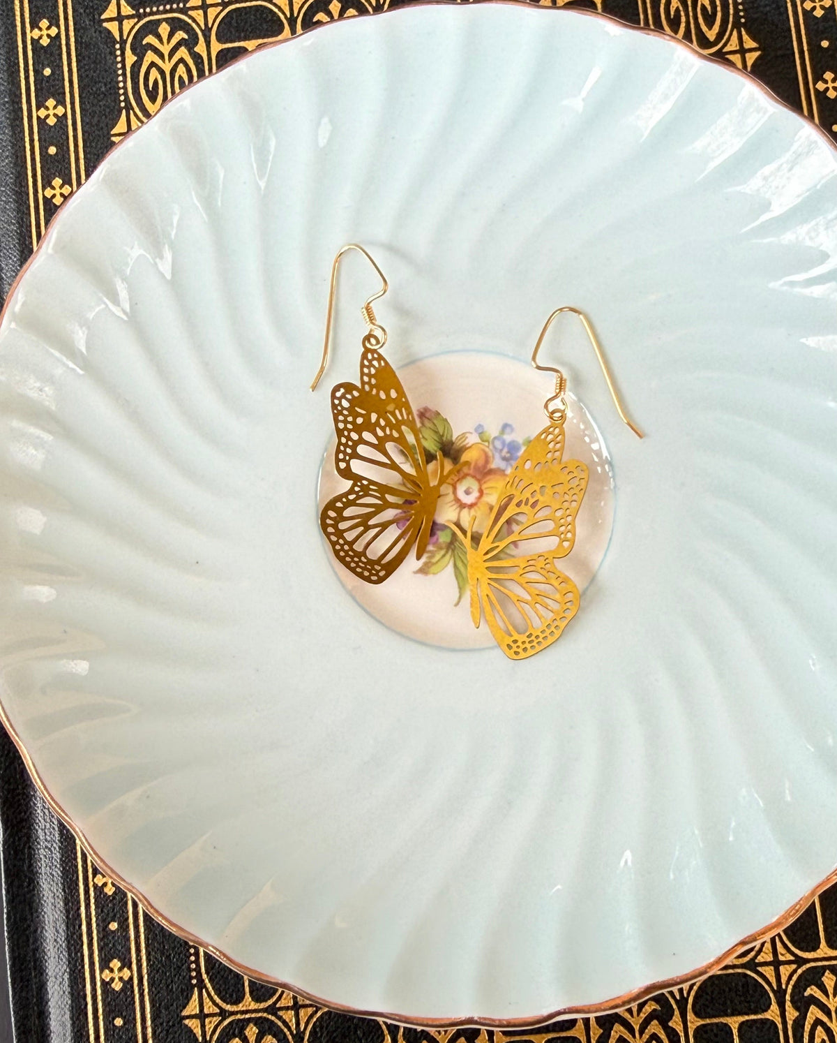 A pair of gold butterfly drop earrings with small floral details, displayed on a white ceramic dish over a black and gold patterned surface.