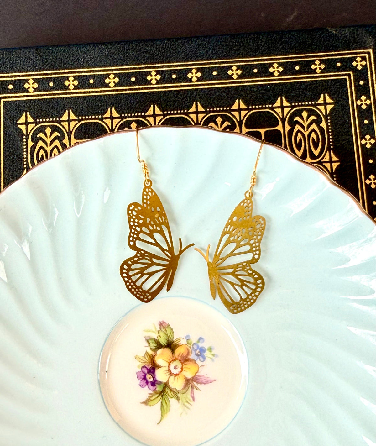A pair of gold butterfly-shaped dangle earrings displayed on a white ceramic plate with a small floral dish underneath, set against a dark patterned background.