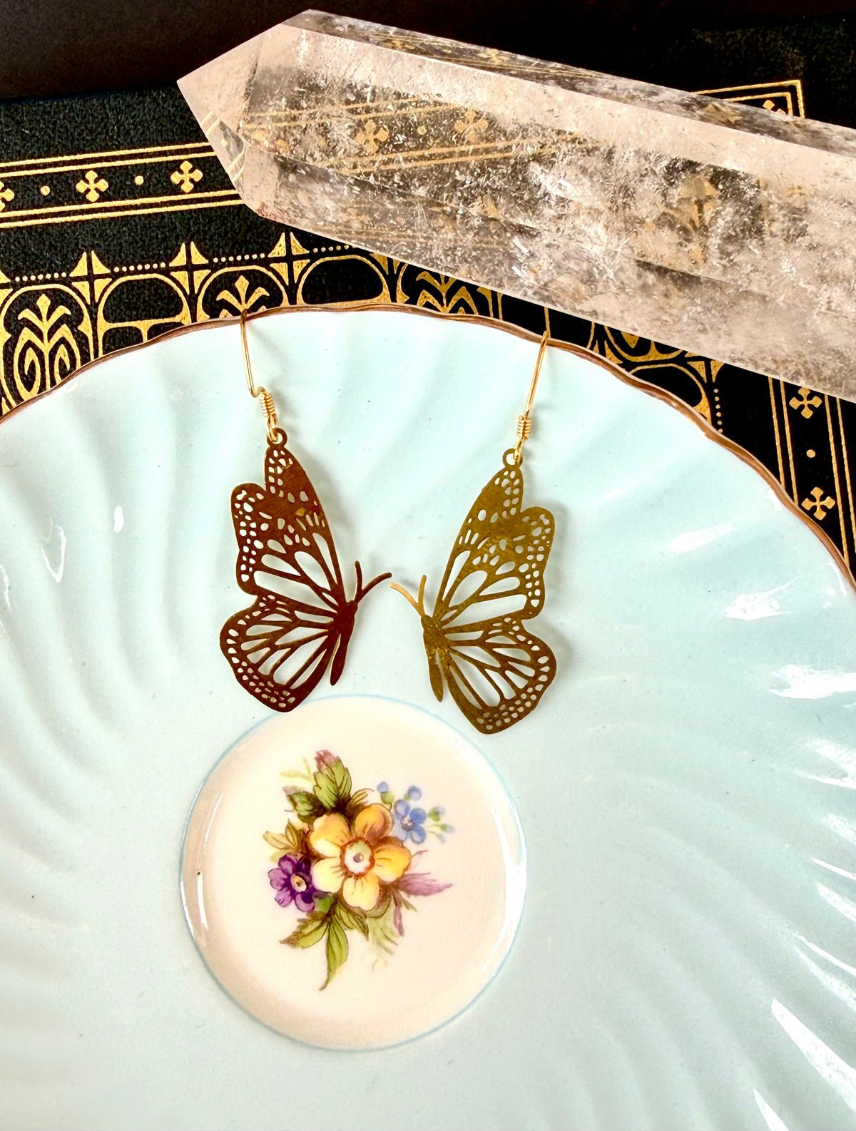 A pair of butterfly-shaped dangling earrings in metallic rose gold and gold tones, displayed on a light blue ceramic plate with a small floral dish and a crystal cluster.