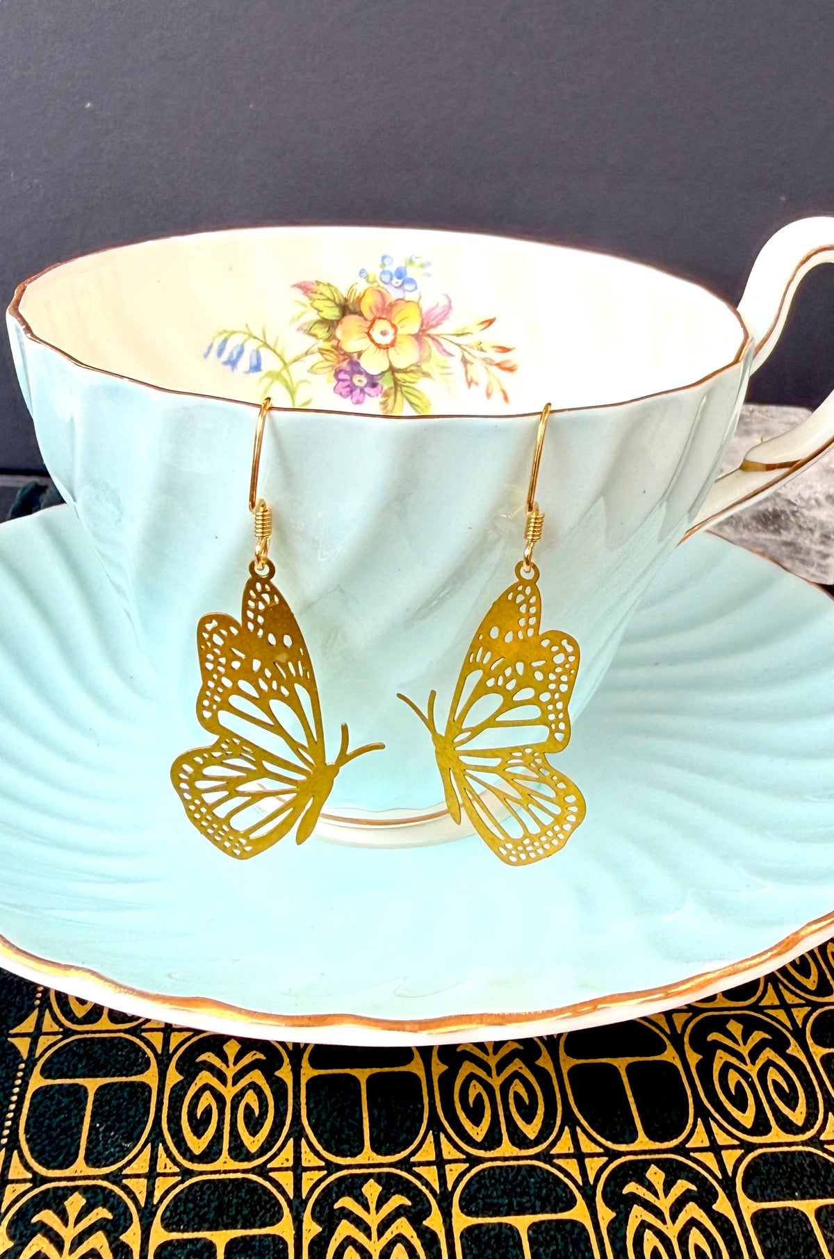 Gold butterfly-shaped dangle earrings hanging from a light blue floral teacup and saucer, set against a dark background with a black and gold patterned surface below.