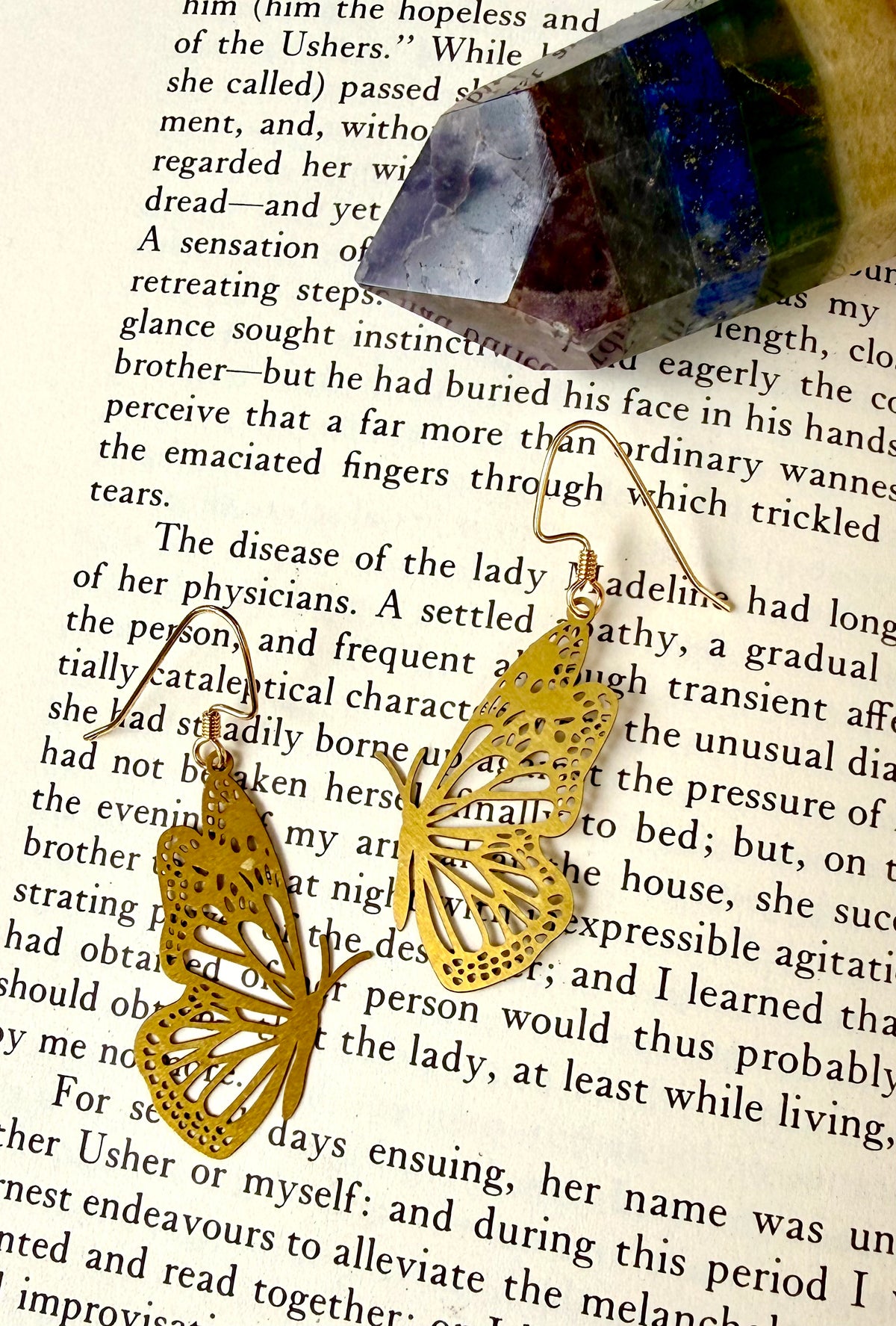A pair of gold butterfly-shaped earrings with intricate cutout detailing, draped over an open book page with a small blue and purple gemstone resting on top.