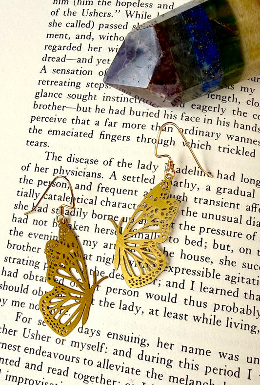 A pair of gold butterfly-shaped earrings with intricate cutout detailing, draped over an open book page with a small blue and purple gemstone resting on top.