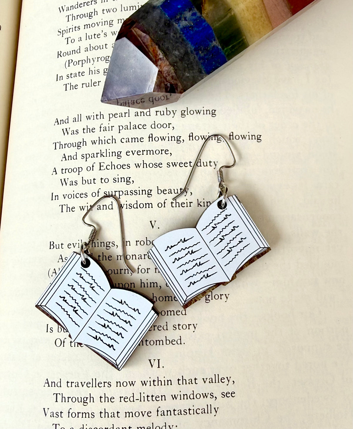 A pair of silver-toned dangle earrings shaped like open books with handwritten text, displayed on an open book page with a colorful glass crystal resting above.