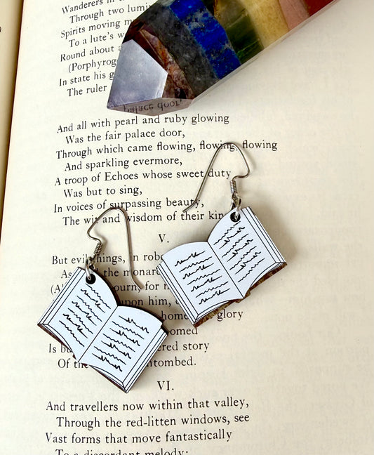 A pair of silver-toned dangle earrings shaped like open books with handwritten text, displayed on an open book page with a colorful glass crystal resting above.