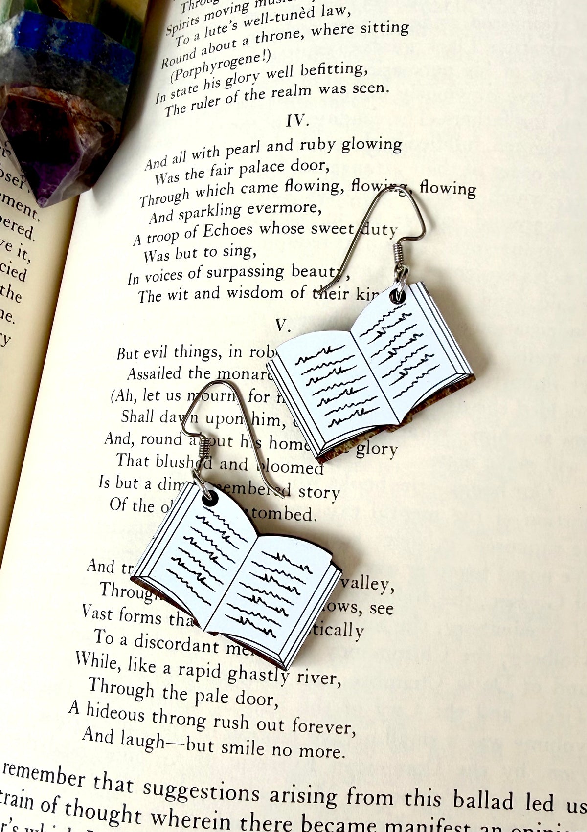 Open book dangle earrings with silver-toned metal hooks and clear acrylic book charms printed with black script, displayed on an open book page.