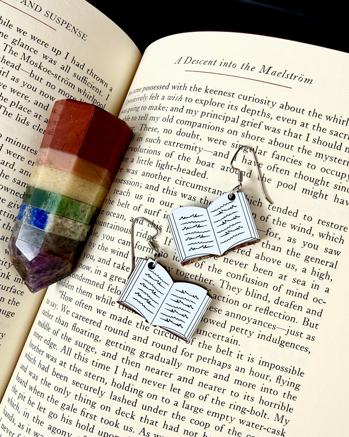 A pair of silver-toned book-shaped earrings with dangling hooks, displayed on an open book next to a small rectangular multicolor crystal, creating a cozy reading-themed composition.