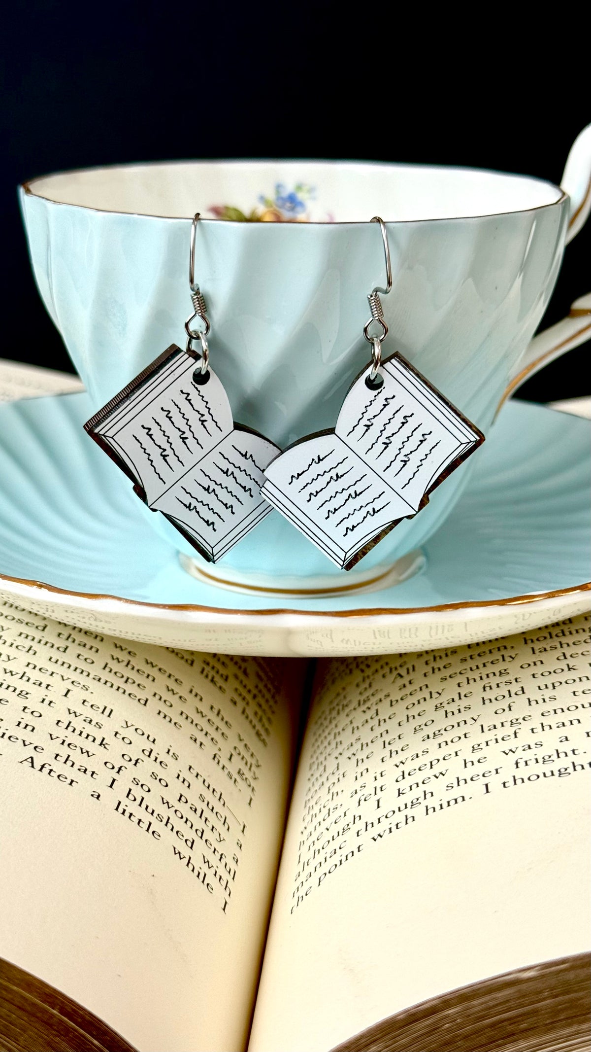 A pair of dangling earrings shaped like open book pages with black script, hanging from silver hooks and displayed in front of a light blue teacup and saucer resting on open book pages.