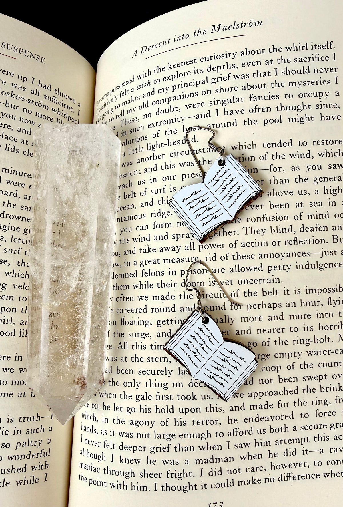 A pair of silver-toned book-shaped earrings with dangling hooks, displayed on an open book next to a small clear crystal, creating a cozy reading-themed composition.