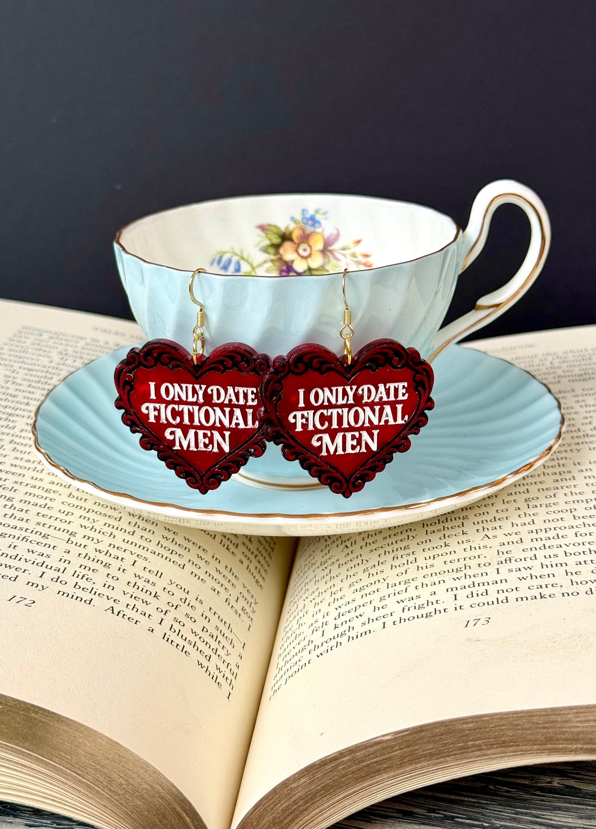 A pair of heart-shaped dangling earrings with red filigree borders and white text reading 'I ONLY DATE FICTIONAL MEN,' hanging in front of a light blue floral teacup and saucer resting on an open book.
