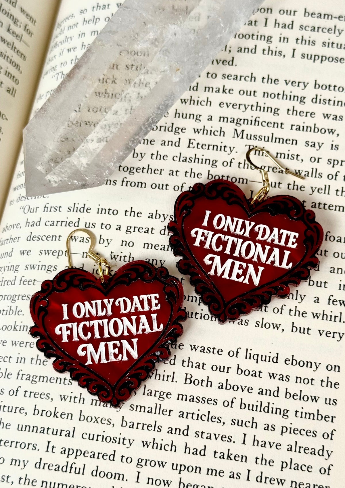 I Only Date Fictional Men red and black heart earrings displayed on an open book beside a clear crystal