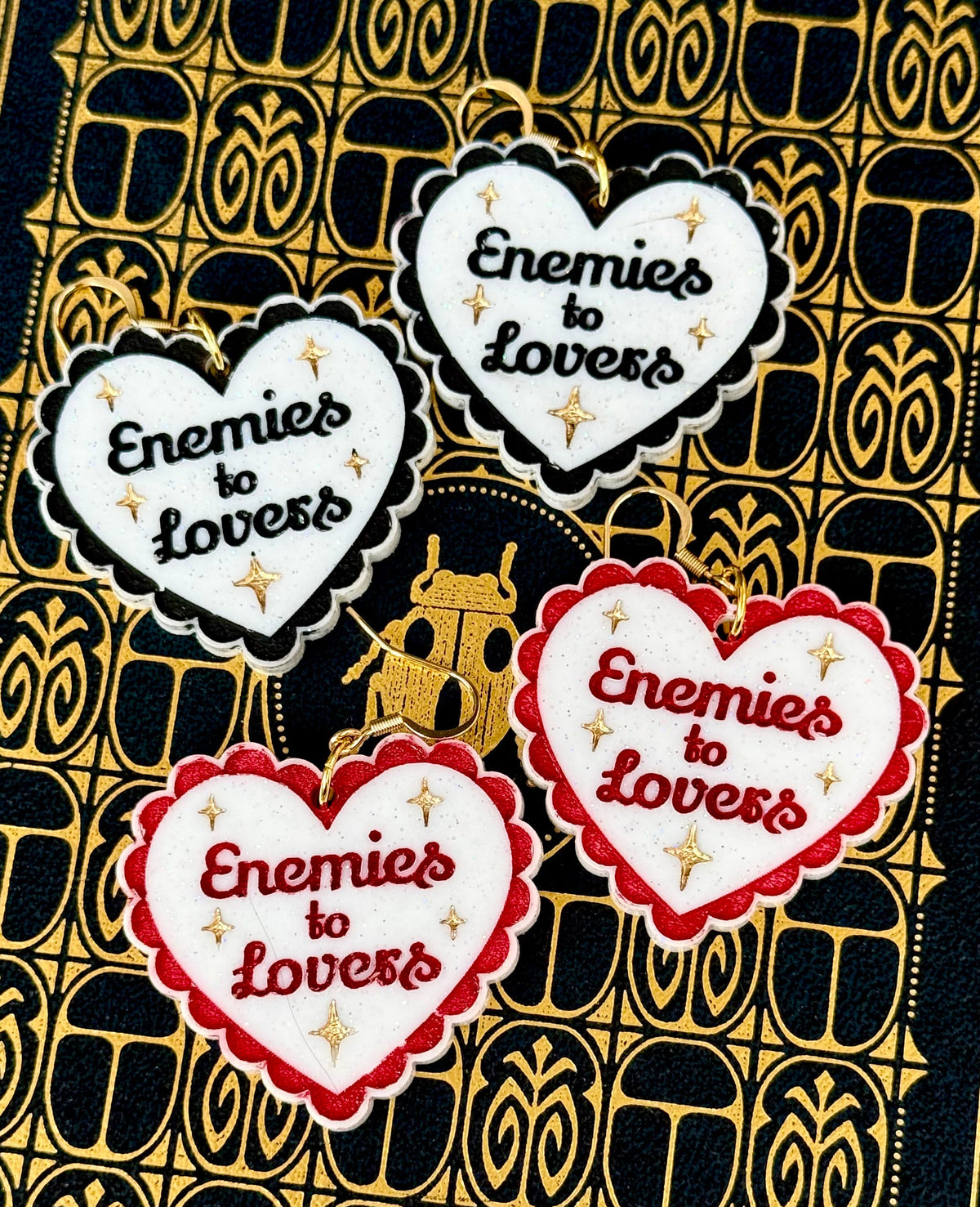 Red and black Enemies to Lovers heart earrings with white glitter centers and gold star details on a black and gold book
