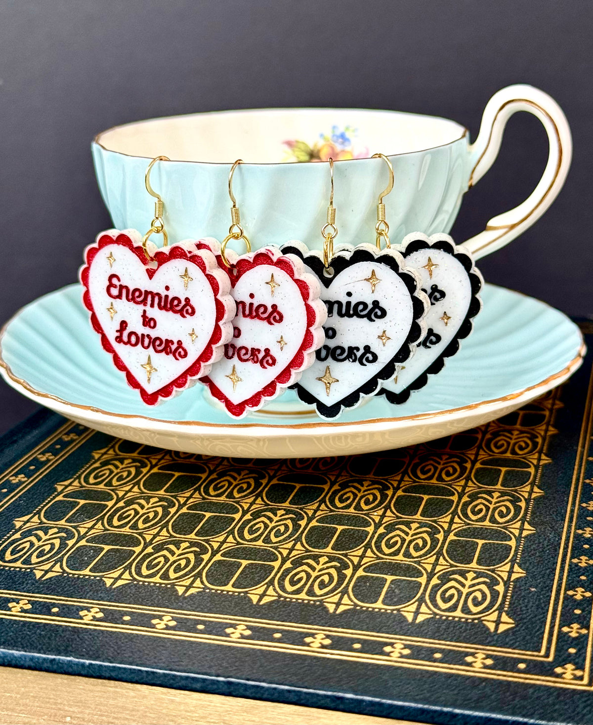 Three heart-shaped dangle earrings with scalloped edges and gold hooks are displayed in front of a light blue teacup and saucer. The earrings feature red, white, and black heart faces with the words 'Enemies to Lovers' in script, accented with small gold stars.