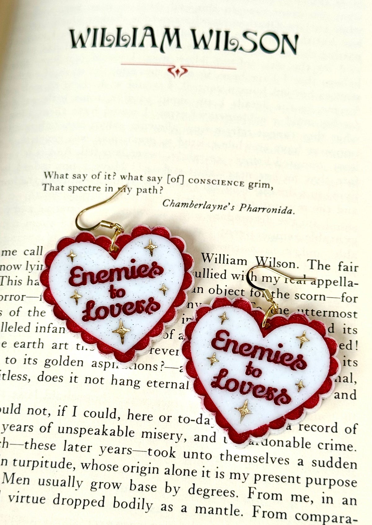 Two heart-shaped enamel pins with red borders and gold lettering reading 'Enemies to Lovers' are displayed on an open book page under the name William Wilson.