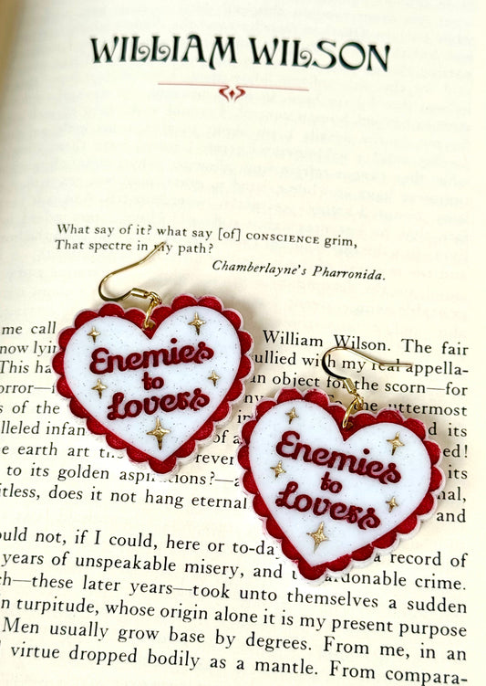 Two heart-shaped enamel pins with red borders and gold lettering reading 'Enemies to Lovers' are displayed on an open book page under the name William Wilson.
