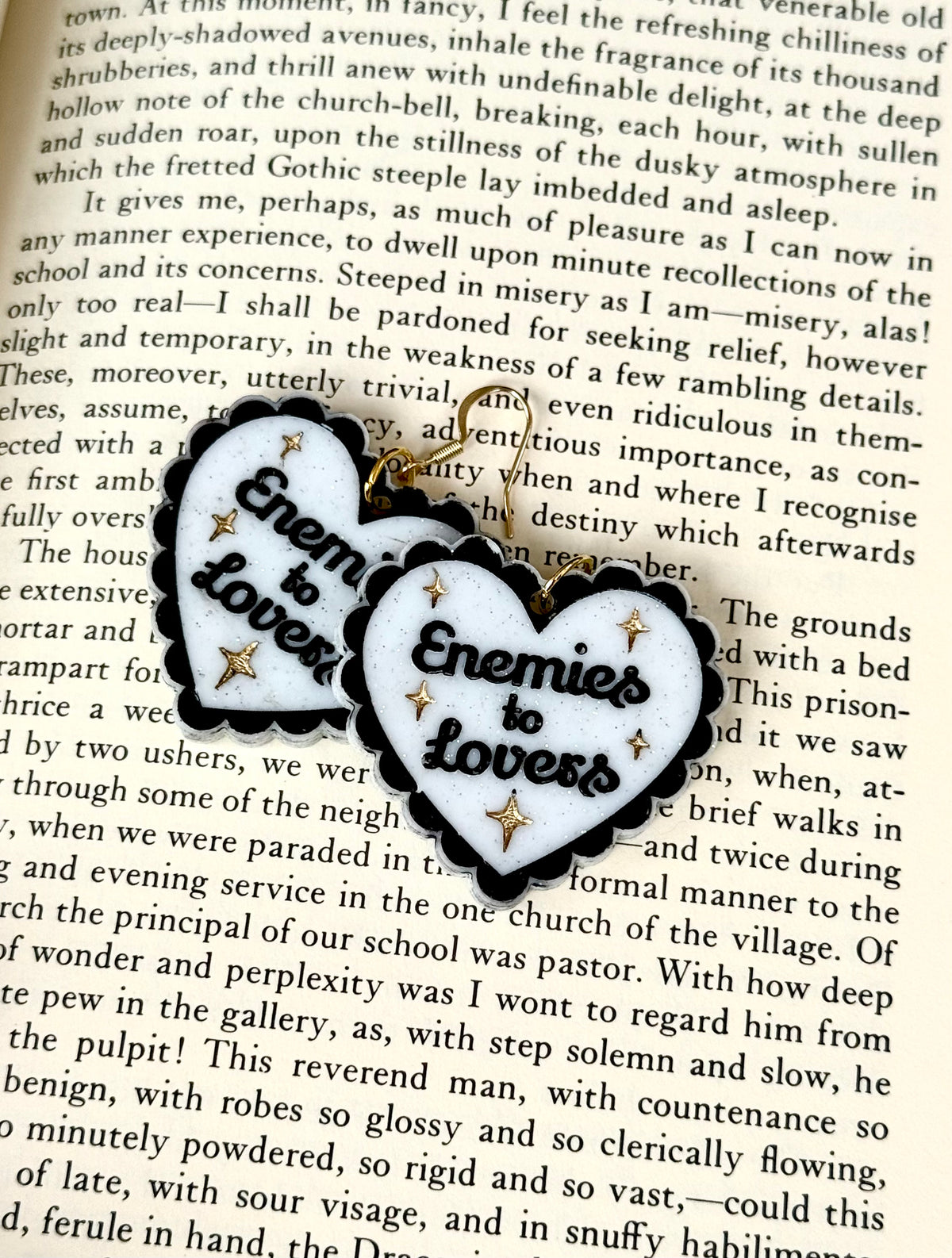 A heart-shaped metal charm with a black and white patterned background and the words 'Enemies to Lovers' in the center, resting on an open book with printed text.