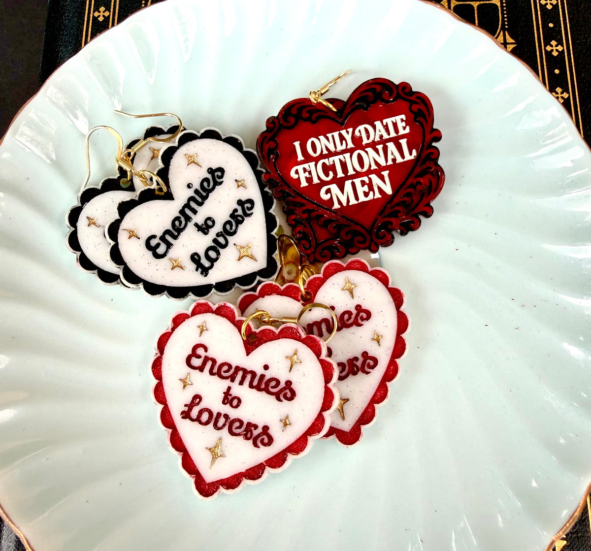 Romance reader earrings including Enemies to Lovers heart earrings and I Only Date Fictional Men earrings displayed on a vintage blue saucer