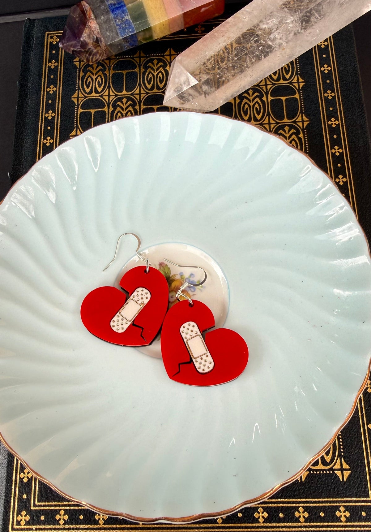 Red heart-shaped dangle earrings with white bandage charms, displayed on a light blue ceramic plate over a black book with gold detailing, with crystals in the background.