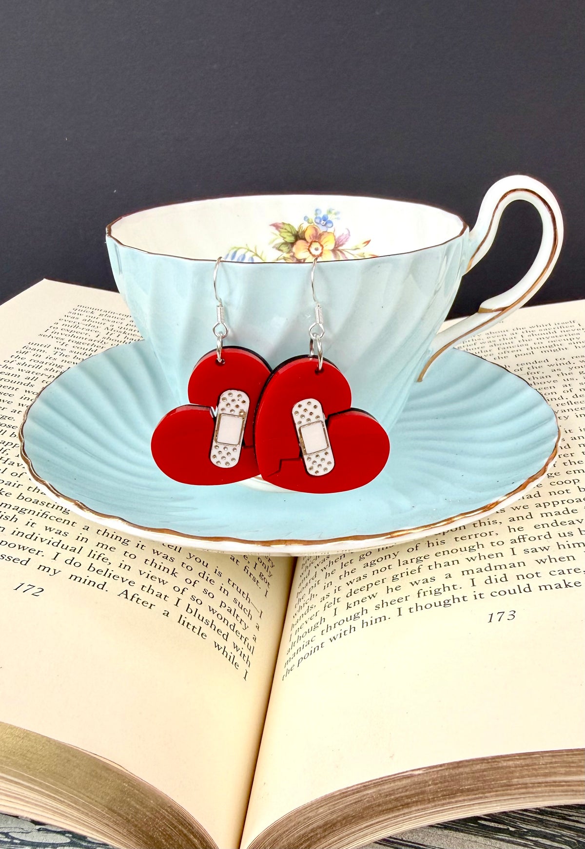 A pair of red heart-shaped dangle earrings with silver hooks and white polka-dot details, displayed on a light blue teacup and saucer resting on an open book.