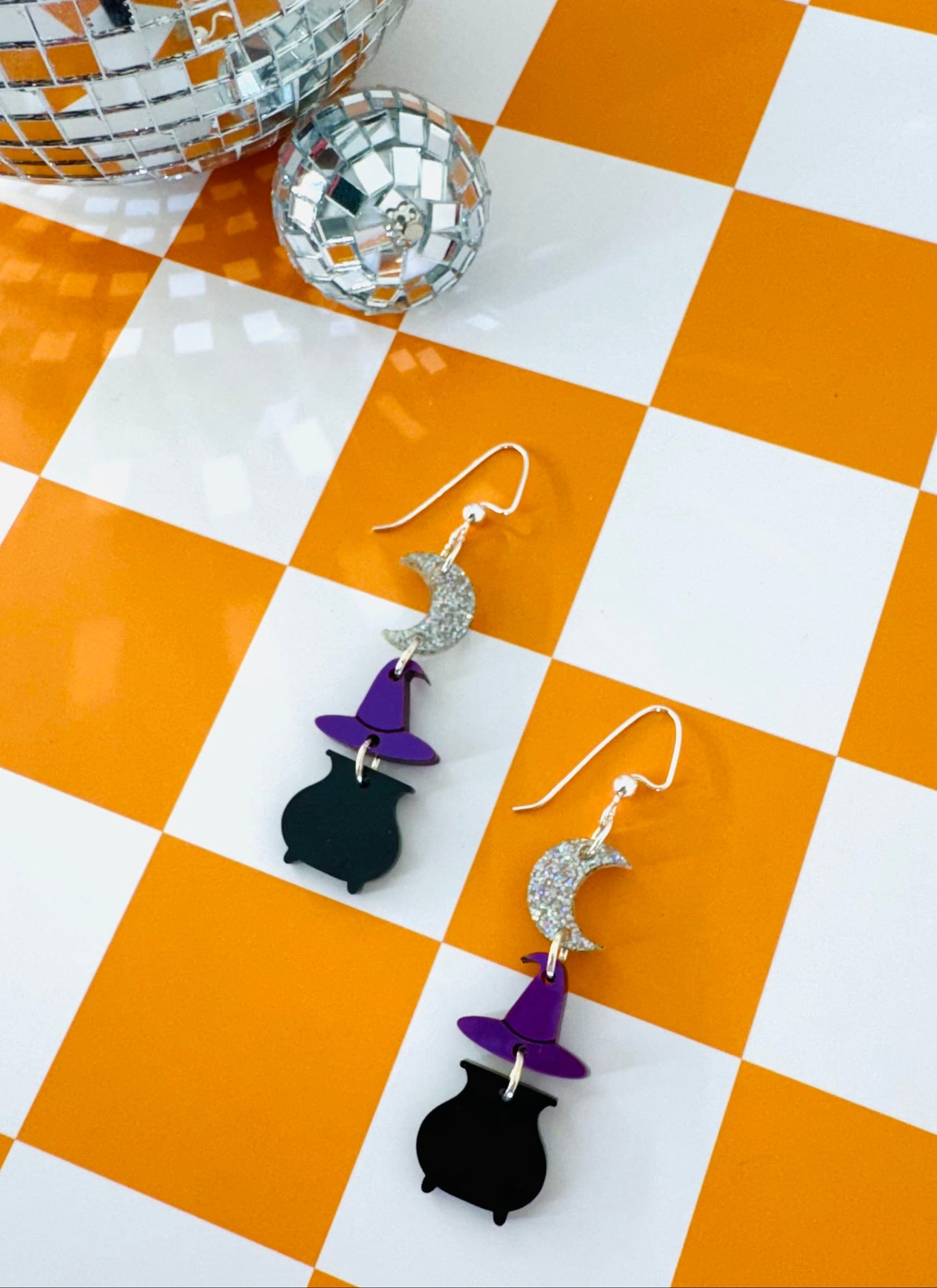 Pair of witch-themed earrings on a checkered orange and white surface with disco balls in the background.