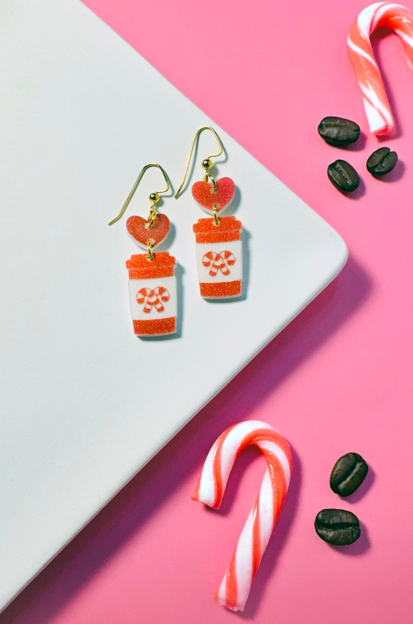 Pair of earrings resembling hot chocolate cups with candy canes and cookies on a pink and white background.