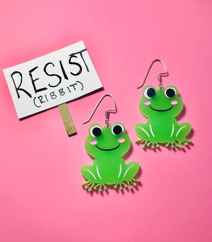 Green frog earrings with a 'RESIST' sign on a pink background