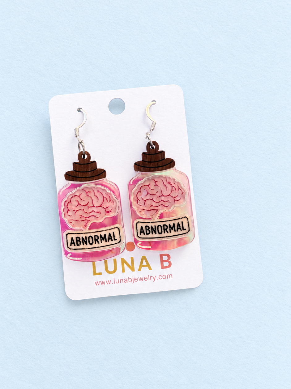 A pair of novelty earrings shaped like pink brain jars with brown lids and a label reading 'ABNORMAL', attached to a white card printed with 'LUNA B' and a website, set against a light blue background.