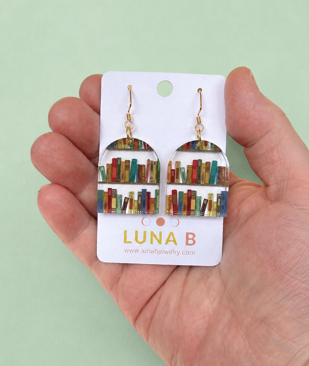 Earrings shaped like bookshelves on a Luna B card held in a hand against a light green background