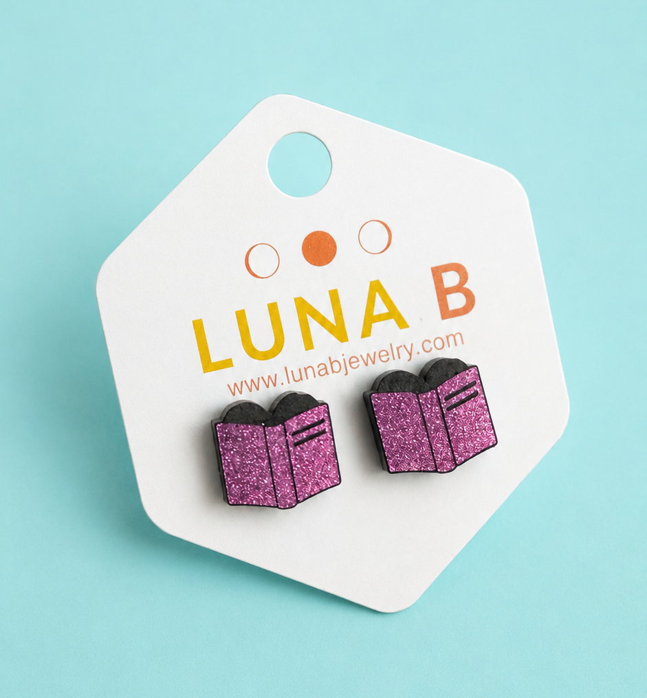 A pair of glittery purple and black book-shaped stud earrings displayed on a white branded backing card against a light blue background.