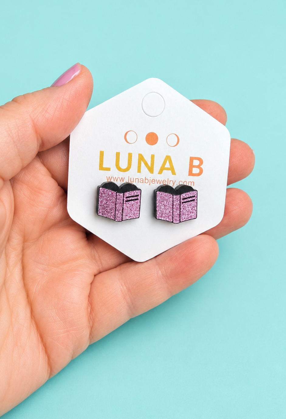 A hand holding a white hexagonal card displaying a pair of small book-shaped stud earrings with glittery purple covers and black spines, against a light aqua background. The card is branded LUNA B.