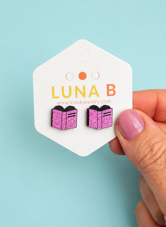A hand with pink nail polish holds a white hexagonal jewelry display card featuring a pair of pink glitter book-shaped stud earrings. The card is branded 'LUNA B' against a solid light blue background.