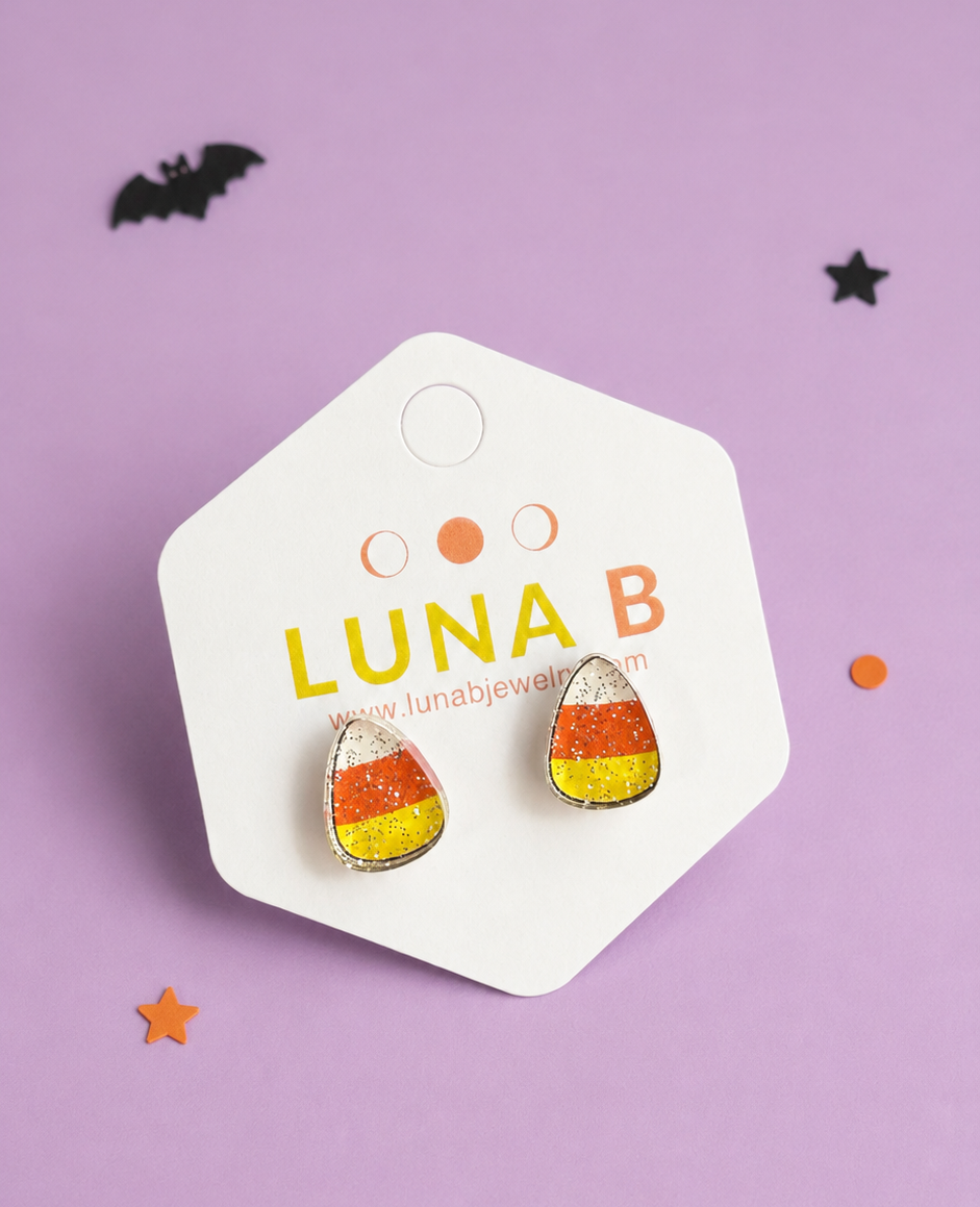 Candy corn themed stud earrings on a white display card, photographed on a lavender background with small black and orange decorative stars and bats.