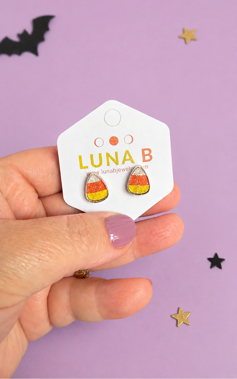 A hand holds a white display card featuring two small candy corn stud earrings with yellow, orange, and white enamel detailing. The card is branded 'LUNA B'.