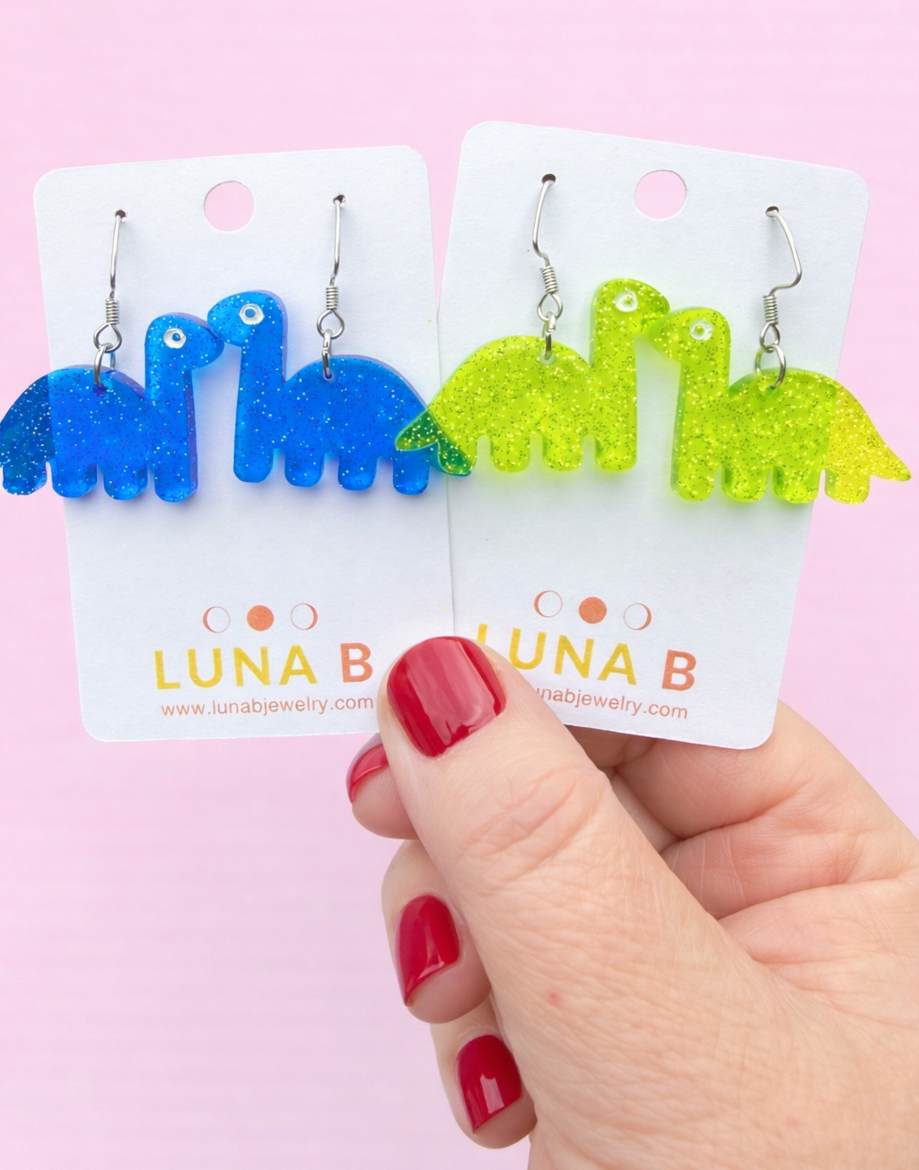 Colorful dinosaur-shaped earrings on Luna B packaging with a hand holding them against a pink background.